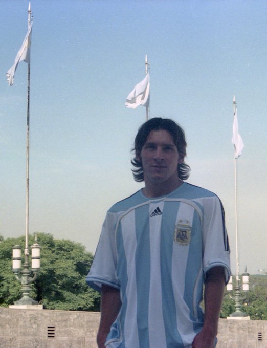 A young man with long dark hair poses in an Argentina national team jersey (featuring the AFA crest and Adidas logo), standing before flagpoles with white flags against a clear blue sky—appearing to be an early-career photo of Lionel Messi, fitting the post's playful wish for his rookie-era trading card to appear in a blockchain pack.
