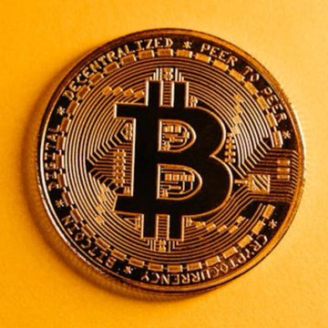 A close-up photograph of a physical Bitcoin coin with its distinctive 'B' logo, resting on a yellow background, featuring the words 'DECENTRALIZED PEER TO PEER' and 'BITCOIN DIGITAL COPPER' inscribed on it.