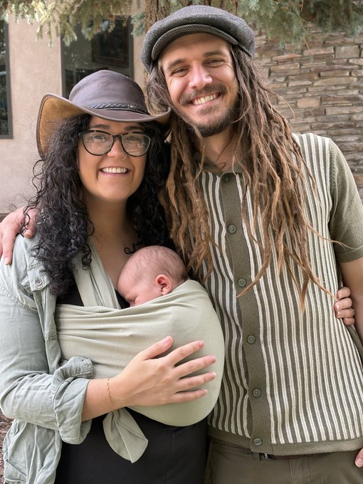 A smiling couple posing outdoors, with the woman wearing glasses and a hat while holding a sleeping baby in a carrier, and the man with dreadlocks wearing a cap and a striped cardigan.