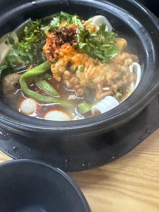 A steaming hot pot of noodle soup topped with cilantro, fried items, and chili in a black clay pot.