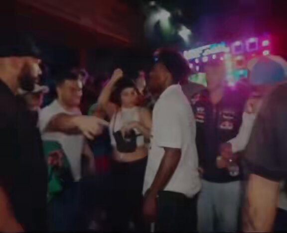 In the image, there are several people gathered together at a party or event, standing around a table with drinks. They appear to be having a good time and enjoying each other's company. One of the individuals stands out, as they are pointing towards someone who is in the center of the room. The person being pointed at has their face partially obscured by something that can be seen hanging on the wall behind them. The overall atmosphere seems lively and social, with people engaging in conversation and sharing drinks from a large communal container placed on the table in front of them.