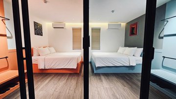 A view into a modern hotel or dormitory room through black door frames, showing two neatly made beds with white linens, wooden floors, and an air conditioning unit mounted on the wall.