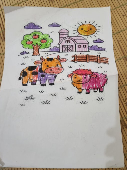 A child's crayon and marker drawing on white paper depicts a smiling sun, an apple tree, a barn, a cow, a pink sheep, and a wooden fence, resting on a bamboo mat.