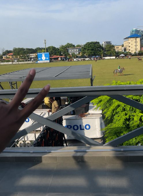 A view from a balcony overlooking a polo field where a match is being played, with a person's hand making a peace sign in the foreground and a 'POLO' sign visible below.