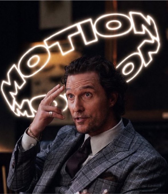 A man in a grey plaid suit and dark tie gestures with his hand near his head in front of a neon sign spelling 'MOTION' in an oval shape.