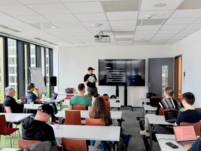 A classroom or workshop setting where a presenter stands at the front speaking to an audience seated at tables with laptops, with a large screen displaying code behind him.