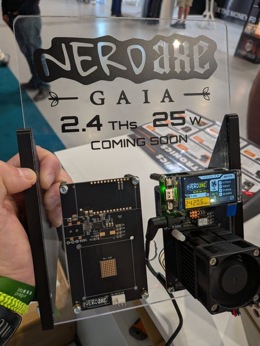 The image features a person holding up a clear plastic case with a black circuit board inside of it. The circuit board has a label that reads " Nerdaxe Gaia - BM 1373 chip 2,4Ths / 25W See it at @BTCPrague Booth". This suggests that the circuit board is related to a project or invention involving Nerda and Gaia chips.