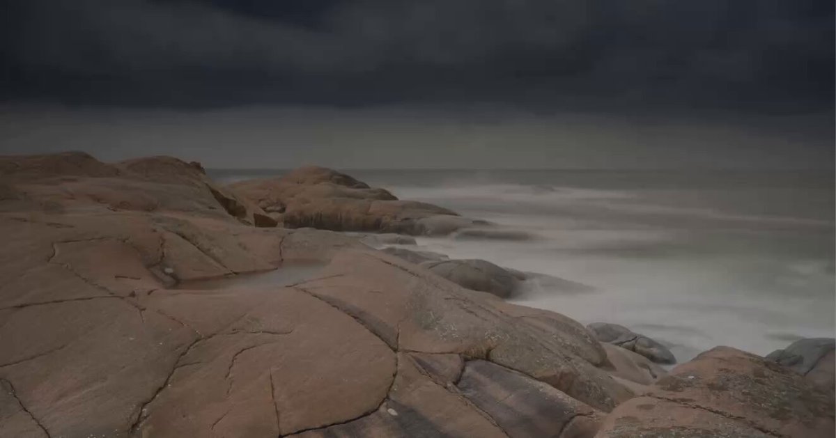 The image shows a rocky shoreline with large rocks and a body of water nearby. The sky above appears dark and cloudy, creating an atmospheric setting for the scene. There are no people or other objects visible in the photo.