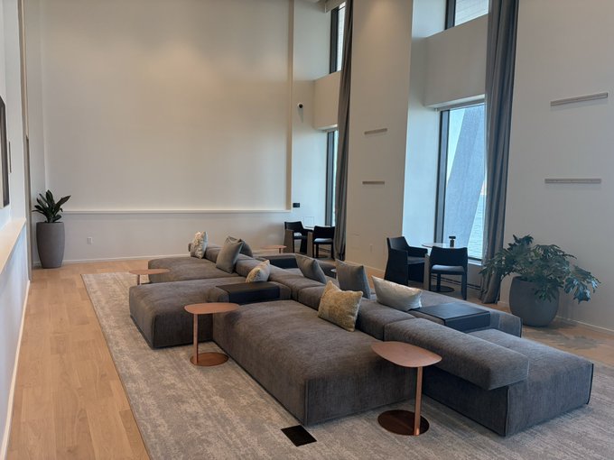 A modern, spacious living room with high ceilings, featuring a large gray sectional sofa with multiple pillows, accompanied by round side tables, a beige area rug, and potted plants near tall windows.