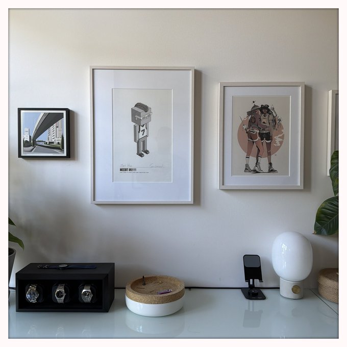 A white desk displaying a watch storage box, a candle, a phone stand, and a white lamp, with three framed artworks hanging on the wall above, including one featuring a robot illustration.