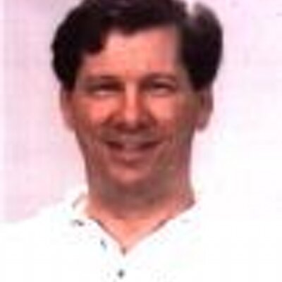 A low-resolution portrait photo shows a smiling man with short brown hair wearing a white shirt.