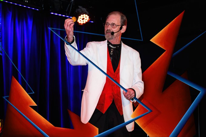A man wearing a headset microphone and a white jacket with a red vest, performing on a stage with blue and orange geometric lighting and structures.