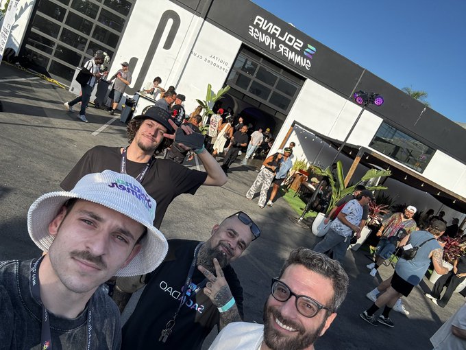 An outdoor selfie of four smiling men standing in front of a building with a 'SUMMER HOUSE' sign and Solara branding, with crowds and event-goers visible in the background on a sunny day.