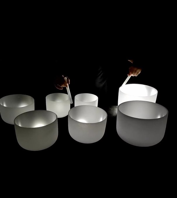 The image features a person holding a large white bowl with their hands while standing next to several other bowls of varying sizes. The person's hands are positioned over these bowls, suggesting that they might be preparing for an activity or performance involving the use of these bowls. The scene is set against a black background, which further emphasizes the focus on the bowls and the person holding them.