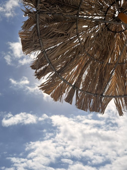 A thatched beach umbrella shown from below against a partly cloudy blue sky. The image serves as a relaxed vacation backdrop, matching the casual, sun-soaked tone of the "GM" greeting and request for Solana wallet addresses.
