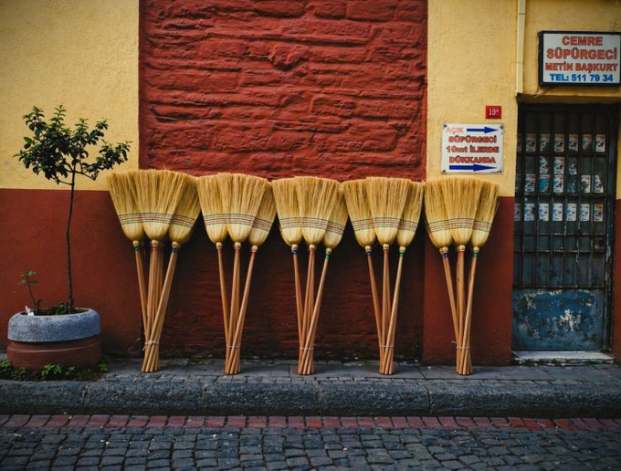 A street scene showing several traditional brooms leaning against a brightly colored red and yellow wall, with a blue door and signage in Turkish (including 'CEMRE SÜPÜRGECİ MİTAT BAKİKURT').