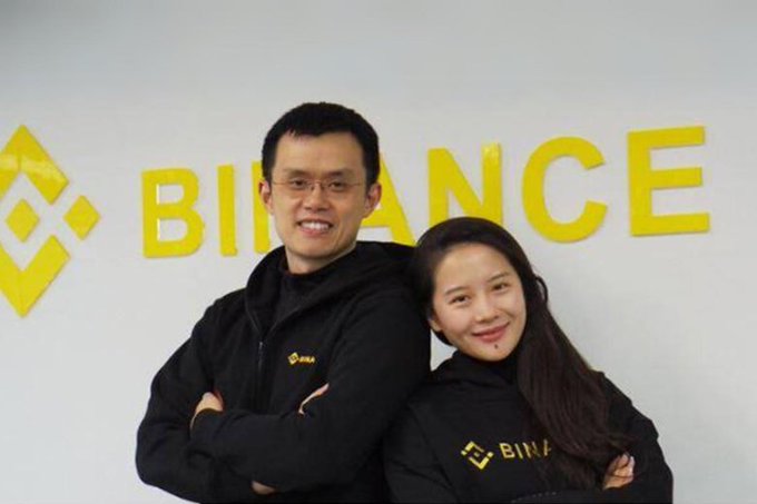A man wearing a black Binance jacket and a woman wearing a black Binance jacket posing together in front of a wall with the yellow Binance logo.