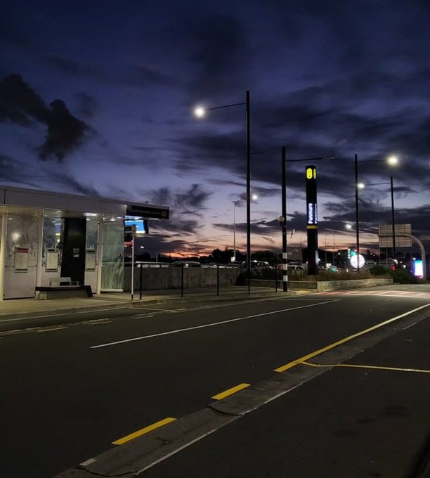 A twilight scene at a bus stop on a city street, with a bus shelter and streetlights under a dramatic purple and orange sky.
