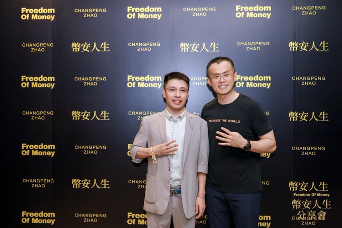 Two men posing in front of a step-and-repeat backdrop featuring "Changpeng Zhao" and "Freedom Of Money" branding, with one wearing a blazer and the other a "CHANGE THE WORLD" t-shirt.
