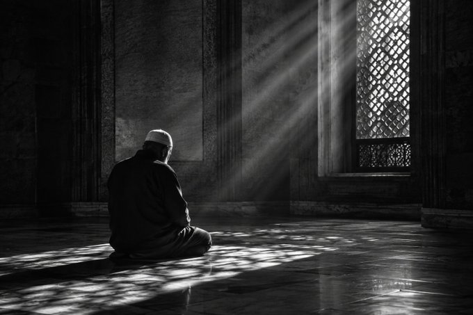 A black and white photograph captures a man wearing a white skullcap, kneeling in prayer inside a dimly lit mosque, with dramatic rays of sunlight streaming through a lattice window.