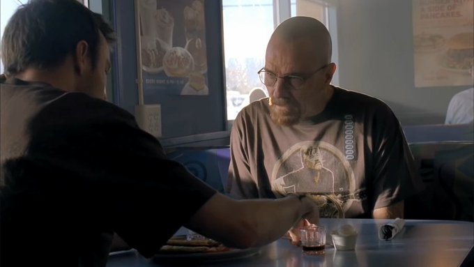 Two men sit at a restaurant booth with a menu for pancakes visible on the wall, one facing away from the camera and another wearing a Breaking Bad-themed t-shirt with a small glass in front of him.