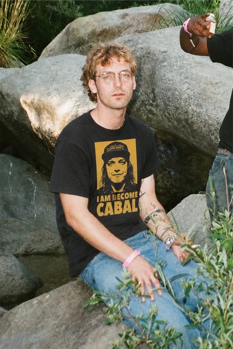 A young man with curly hair and glasses sits on rocks, wearing a black "I AM LECOME CABAL" t-shirt featuring a stylized portrait, with visible tattoos on his forearms. The "Cabal" shirt text ties into the "Saving Ethereum" post, likely referencing the infamous 2016 DAO hack cabal group.