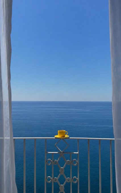 A bright yellow coffee cup sits on a white wrought-iron balcony railing, framed by flowing sheer curtains overlooking a calm blue sea under a clear sky—a peaceful morning scene that perfectly complements the warm "Good morning family" greeting.