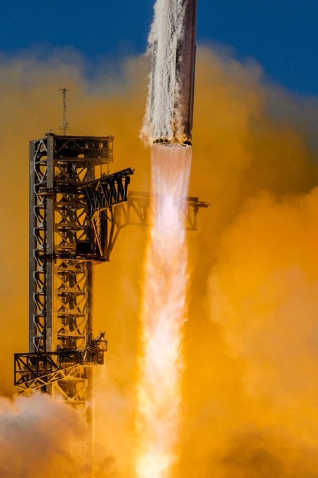 A dramatic photograph of a rocket launching from a launch tower, with bright orange flames and white smoke billowing out as it ascends against a blue sky.