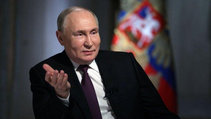 Vladimir Putin is shown in a formal interview setting, gesturing with his hand while speaking, with the Russian coat of arms visible in the background, providing visual context for the post about Russia officially registering its first crypto exchanges and approving Sberbank for crypto custody services.