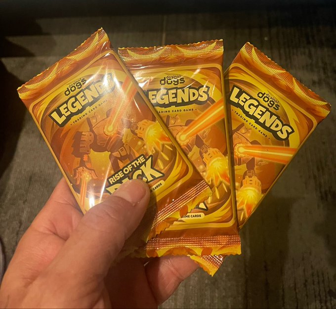A hand holding three sealed orange foil packs of 'Topps Dogs of the Worlds Legends Trading Card Game' featuring a robot illustration and text reading 'Rise of the'.