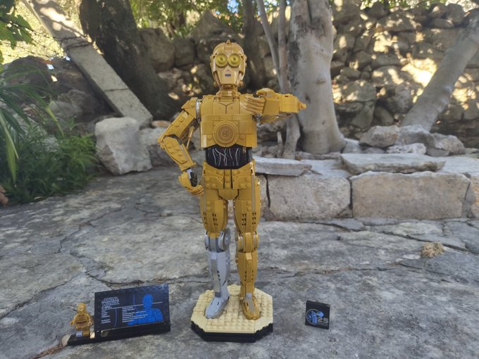 A LEGO C-3PO minifigure display stands on a stone surface in a garden setting, with small information placards at its base.
