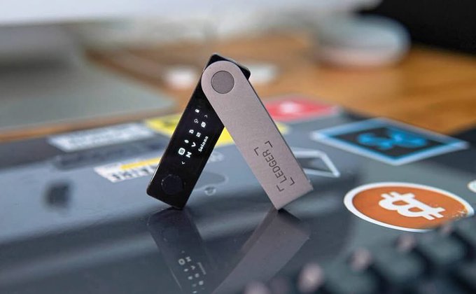 A close-up of a Ledger cryptocurrency hardware wallet standing upright on a laptop covered with crypto-related stickers, including a Bitcoin logo.