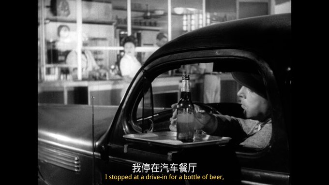 The image shows a man sitting inside an old car, holding a beer bottle up to his mouth while driving. The scene takes place in front of a restaurant or diner, with people visible through the window. There are also other people present in the background who appear to be dining at the establishment.