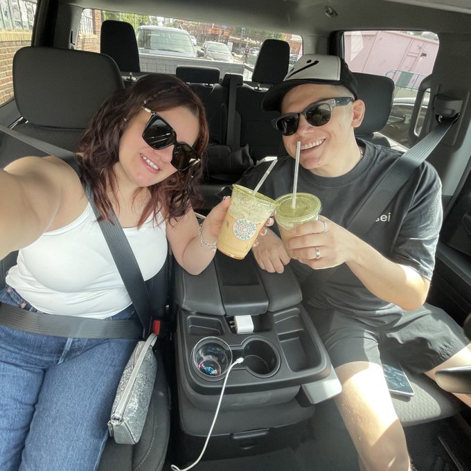 A selfie of a man and woman sitting in a vehicle's center console, smiling and toasting with iced Starbucks drinks while wearing sunglasses.