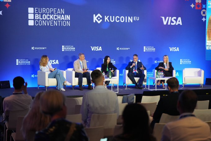 A panel discussion at the European Blockchain Convention with speakers seated on stage in front of a blue backdrop displaying logos for European Blockchain Convention, KuCoin EU, and Visa, with an audience visible in the foreground.