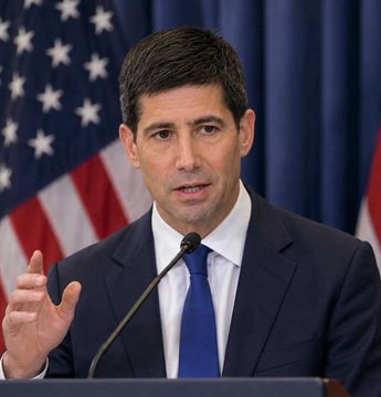 A man in a dark suit and blue tie speaks at a podium in front of an American flag, gesturing with his hand, conveying a sense of authority and urgency in his message.