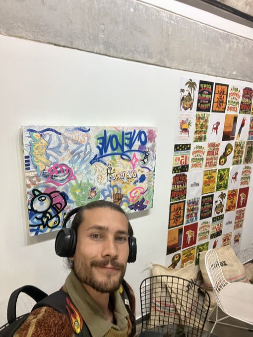 A man wearing headphones and a backpack takes a selfie in front of colorful abstract art and a poster featuring 'HAWAII' designs.