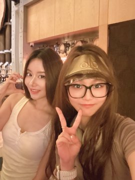 Two young women pose together for a photo; one wears a white tank top while the other wears a brown shirt, glasses, and a cap, both making peace signs.