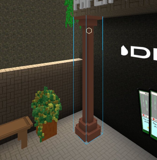 A voxel-style 3D scene shows a brown pillar in an enclosed area with brick walls, a small bench, a plant, and a digital sign, with a hexagon marker on the pillar.