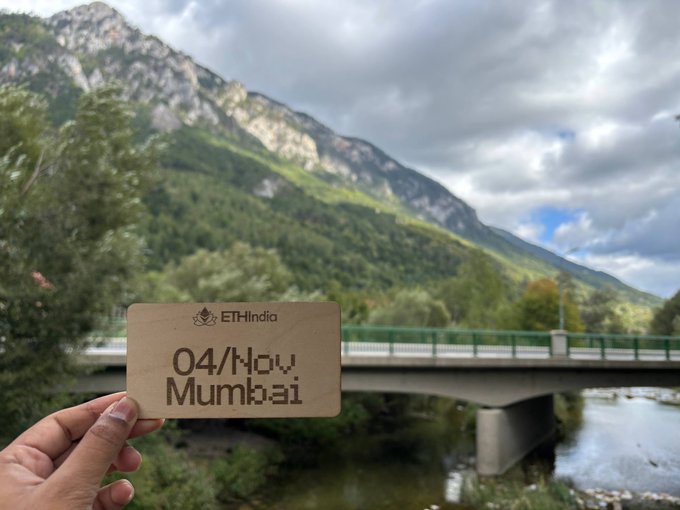 A hand holds an "ETHIndia" ticket dated "04/Nov" and "Mumbai" against a scenic backdrop of mountains, a bridge, and a river.