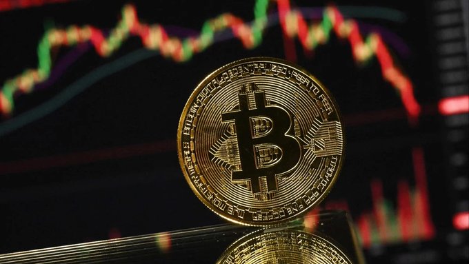 Close-up photograph of a gold Bitcoin commemorative coin with the 'B' symbol, set against a background showing a candlestick price chart with green and red bars.