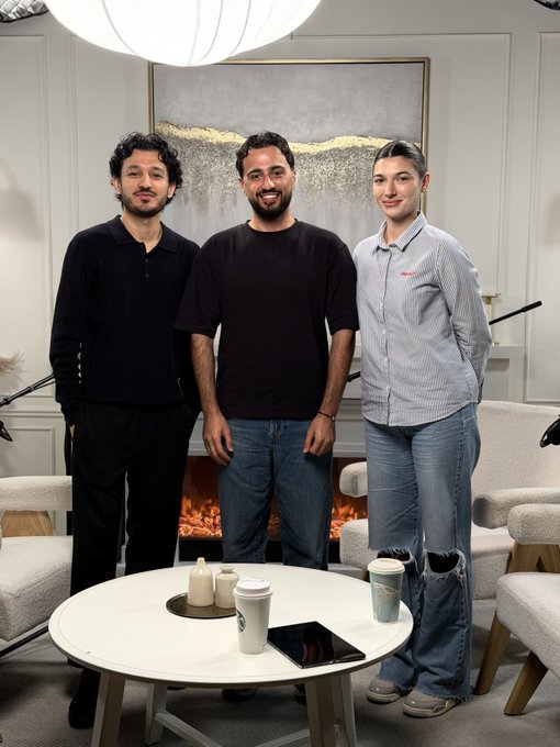 Three people stand smiling in an elegant, modern living room with white walls, a fireplace, and abstract artwork, gathered around a coffee table with coffee cups. The post indicates this is a promotional photo featuring the team behind @Outcomexyz, likely captured during or before an interview or podcast conversation.