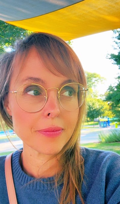 A selfie of a young woman with glasses and a blue sweater standing under a yellow canopy with greenery behind her.