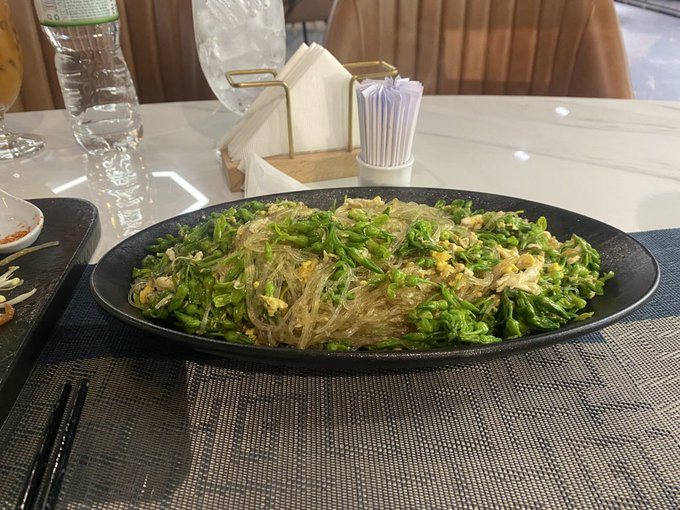 A plate of stir-fried glass noodles with green vegetables (resembling fried water spinach or bean sprouts) served on a black dish at a restaurant table with water bottles and chopsticks.