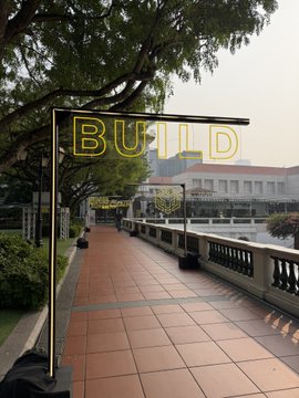 An outdoor walkway with a neon yellow 'BUILD' sign overhead and trees on the left side.