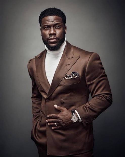 A man in a brown double-breasted suit with a white turtleneck and pocket square poses for a portrait against a gray background, wearing a wristwatch.