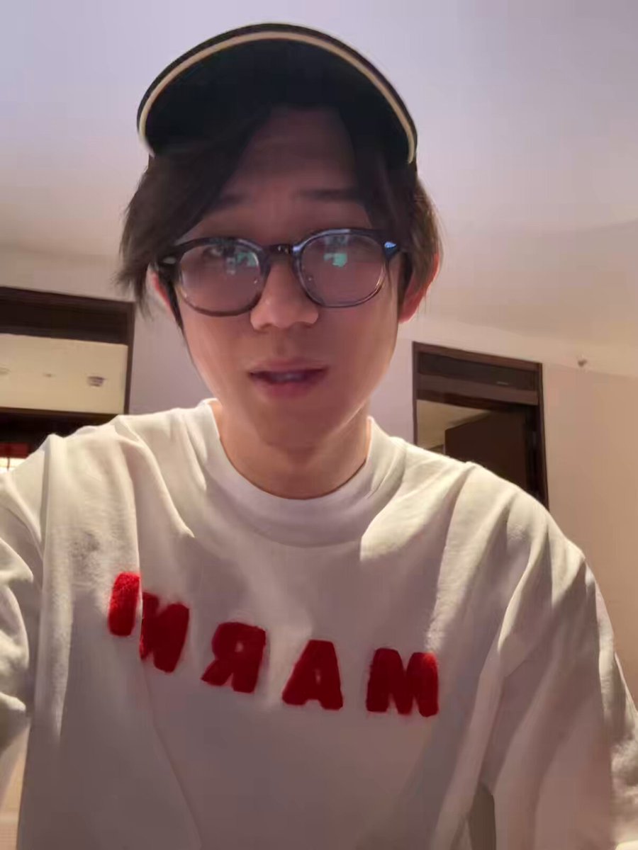 The image features a young man wearing glasses and a white shirt with red letters that say "MAR". He appears to be posing for a photo, possibly for a social media platform or an advertisement. The background of the image shows a wall, which could be part of a room in someone's house or another setting where he is taking the picture.