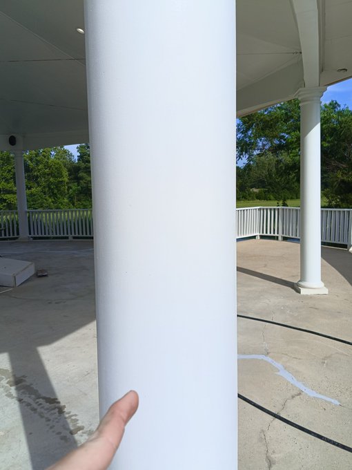 A hand touches a freshly painted white column of a gazebo, with the surrounding structure and outdoor scenery visible. The post proudly displays the recent paint job, fitting the caption "I painted this post. Am artist now."
