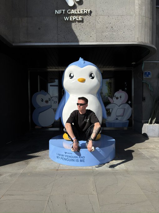 A man crouching in front of a large penguin statue outside the 'NFT GALLERY WEPLE', which reads 'I AM MY PENGUIN AND MY PENGUIN IS ME'.