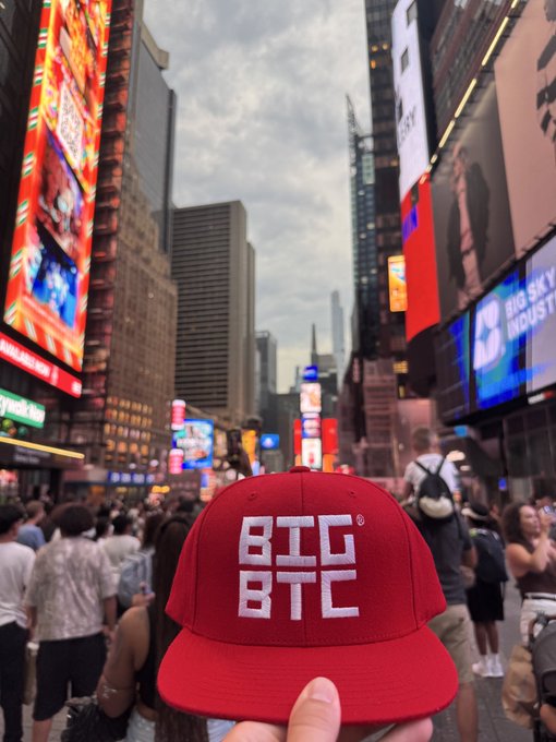 A hand holds up a red baseball cap with "BTC BTC" embroidered in white lettering against the backdrop of a bustling city street with bright billboards.